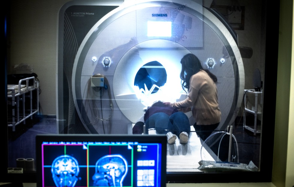 A patient lies on a sliding table being positioned into a large MRI scanner while a technician stands beside them, adjusting their placement. The scene is viewed through a window from a control room, where computer monitors display brain scan images in the foreground.