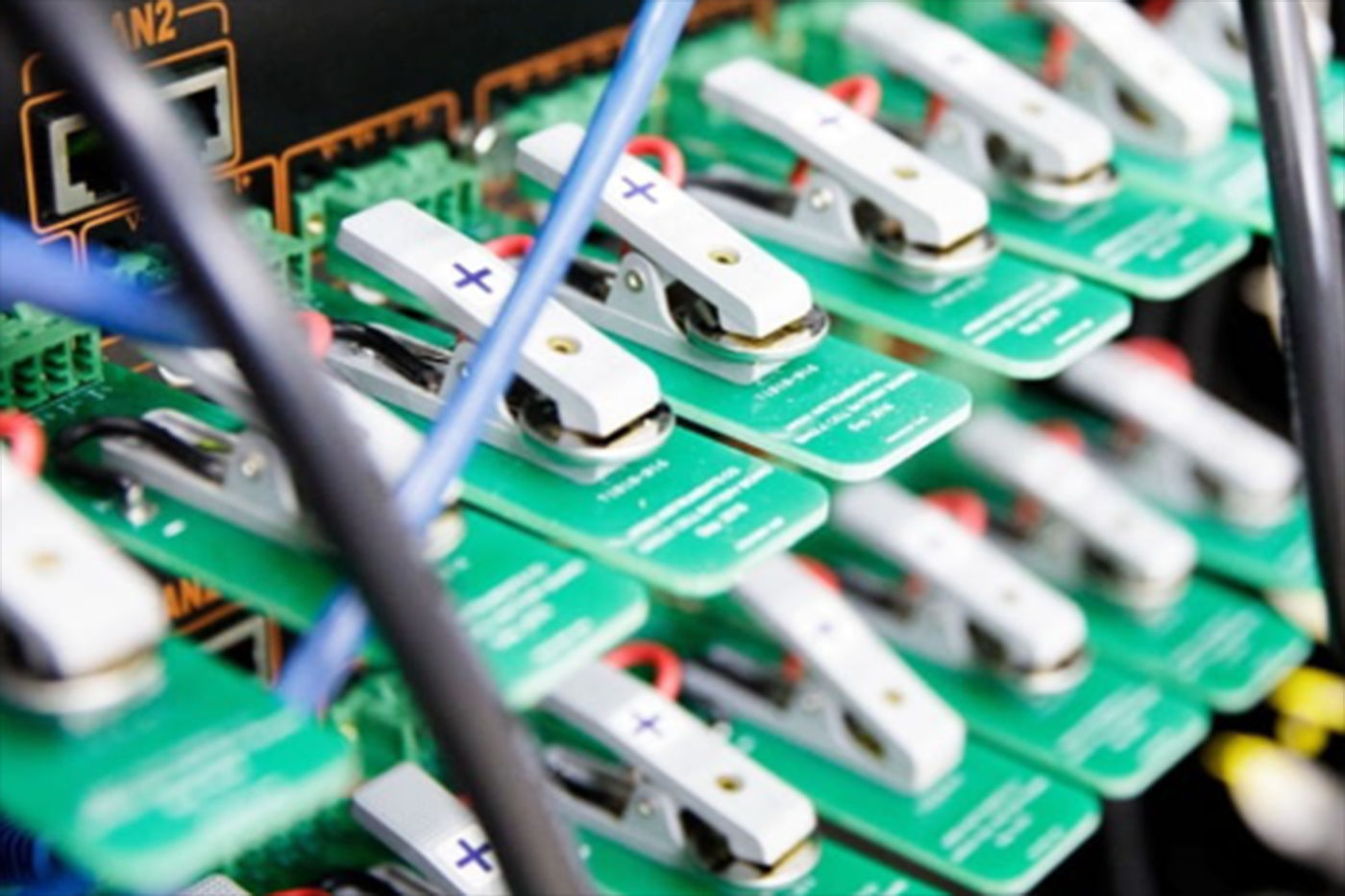 Close-up of multiple green circuit boards or battery modules connected in a rack with white clip-like components and colored wiring, suggesting electronic testing or charging equipment in a data center or lab environment.
